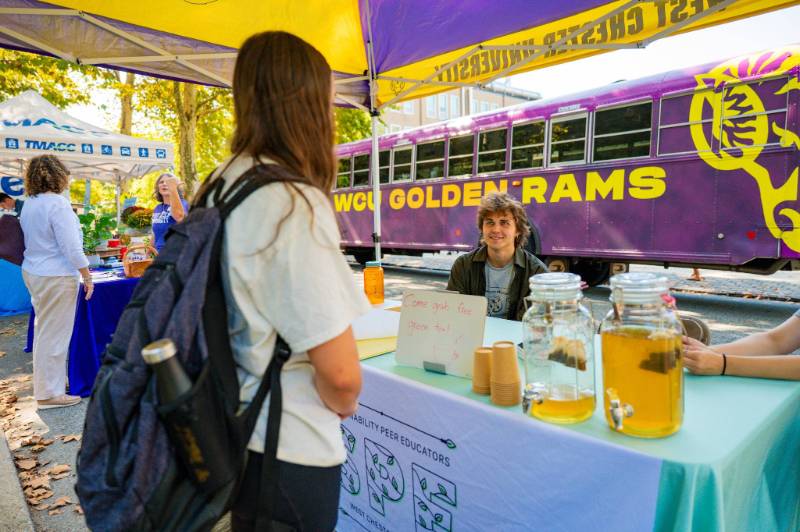 A student sits behind a table offering free green tea as part of Park(ing) Day 2025. The table is set up in a parking space, and other installations can be seen in the background, as can a WCU shuttle bus driving by.
