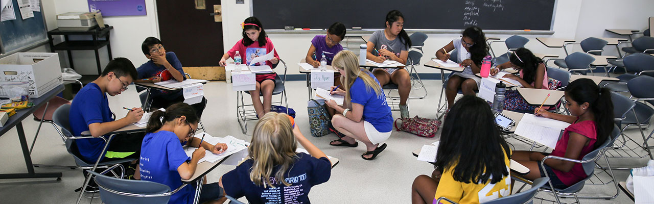 Students sitting in circle of desks while teacher stands in middle instructing