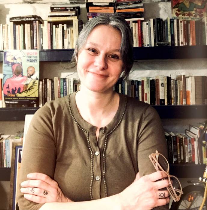 individual with grey-ish hard, light skinn, anda a brown button-up blouse in front of a full bookshelf