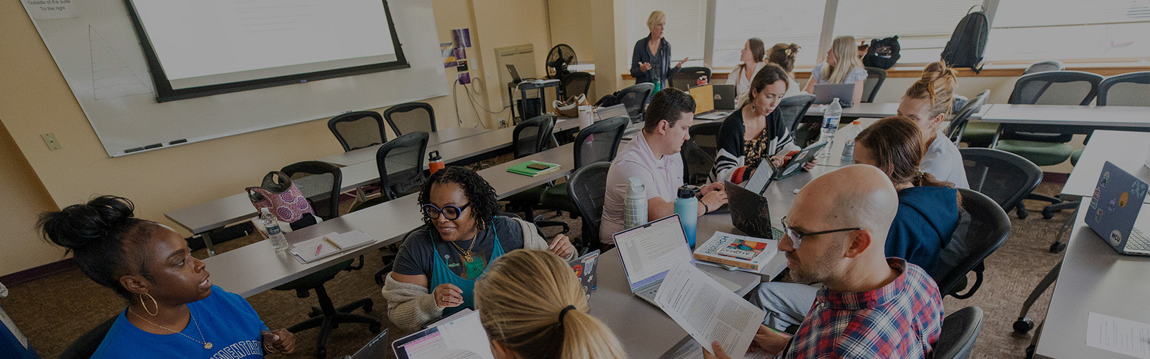 A group of people sit around tables in a classroom or meeting room, working on laptops and discussing papers.